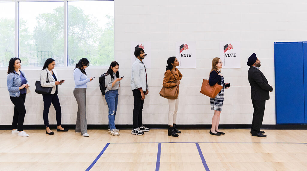 A multiracial group of people, many with smart phones, line up along a wall in a school gymnasium in order to vote