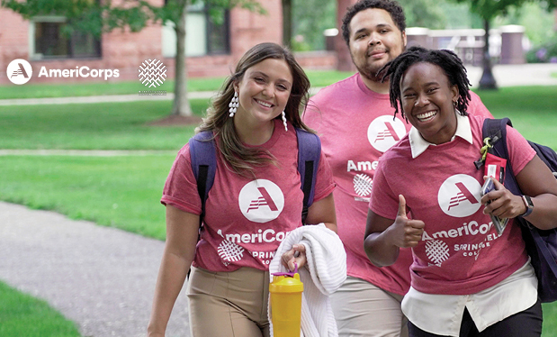 three students wearing AmeriCorps t-shirts are walking across campus giving thumbs up