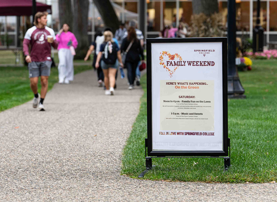 a lawn sign with family weekend events on it with groups of people walking across campus behind the sign
